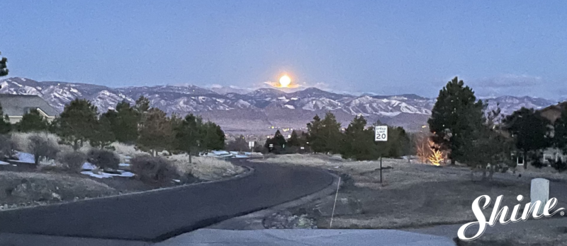 Moonrise illuminates snow-covered mountains and a winding suburban road at dusk.