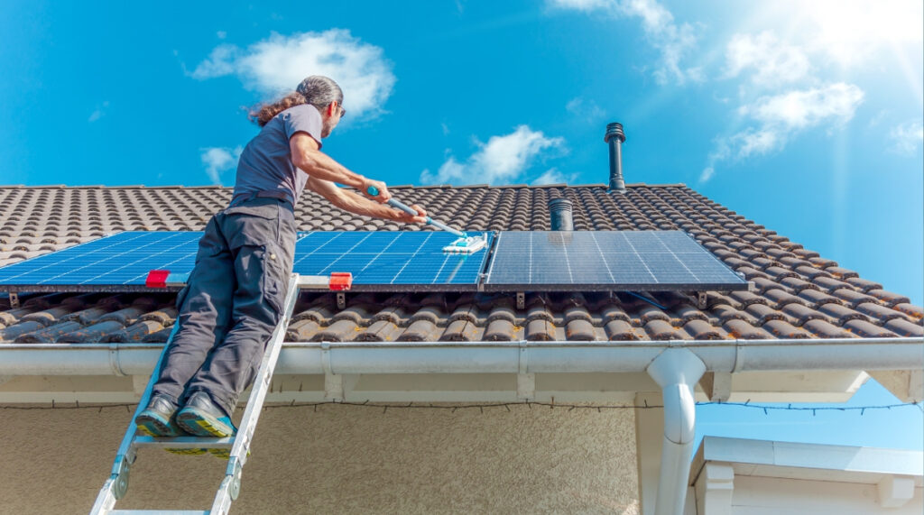 A man on a ladder cleans solar panels on a residential tiled roof under a bright