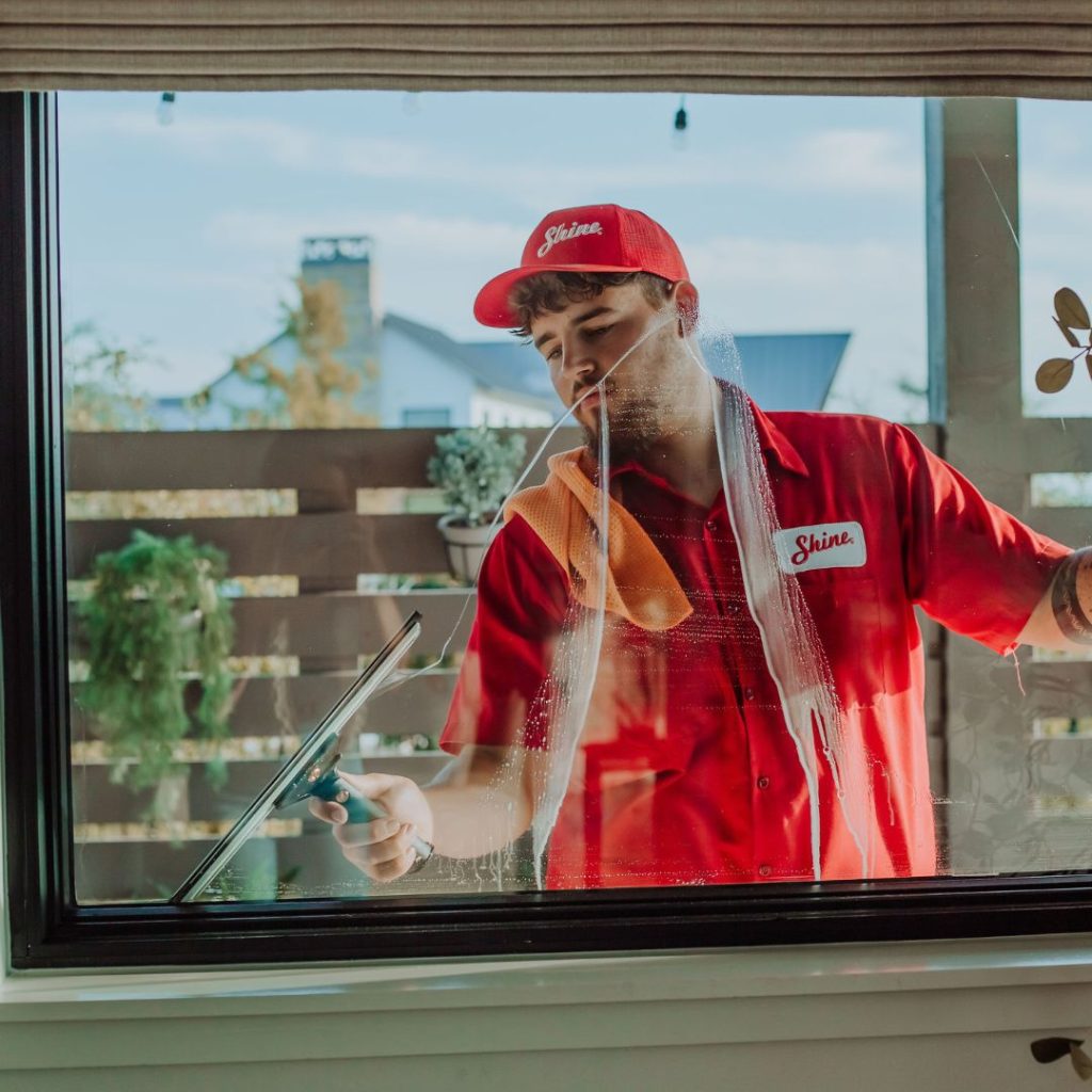 A man in a red "Shine" uniform expertly cleans a large window with