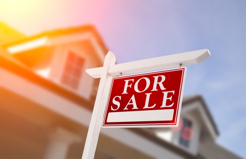 A vibrant red 'FOR SALE' sign is displayed on a white post with a blurred house