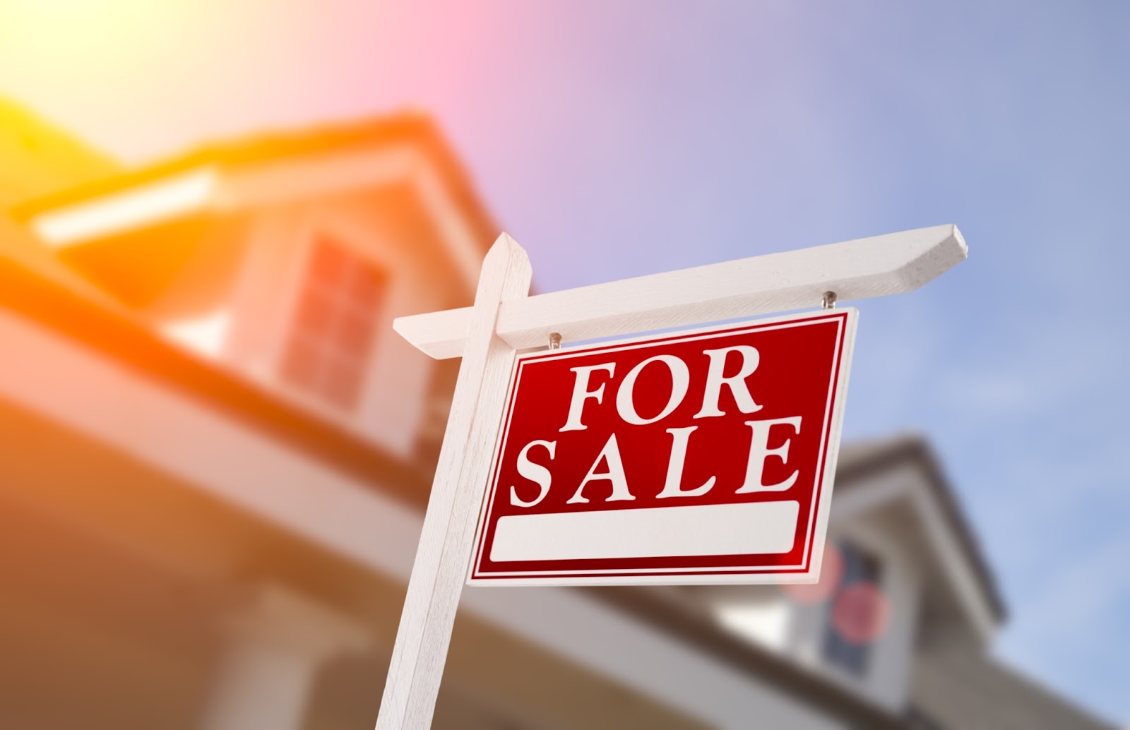 A vibrant red 'FOR SALE' sign is displayed on a white post with a blurred house