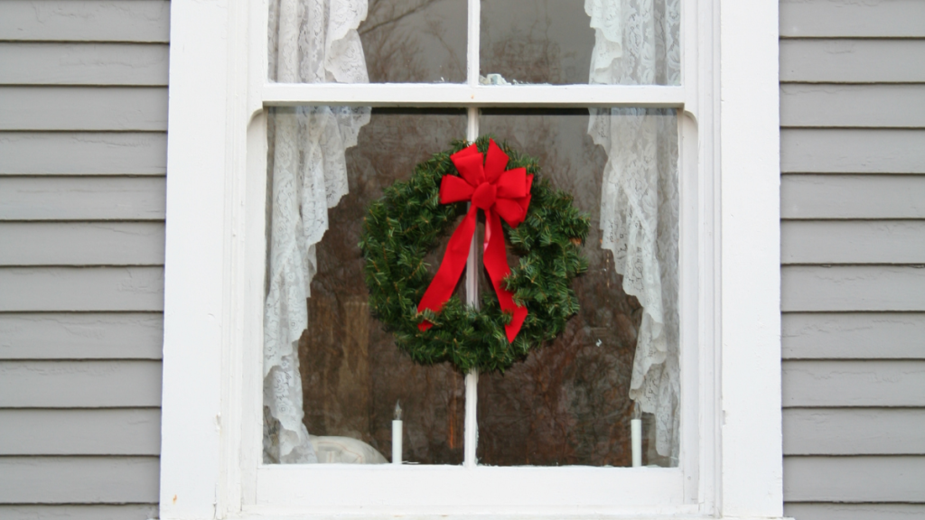 A festive green Christmas wreath with a large red bow hangs in a white-framed window with lace