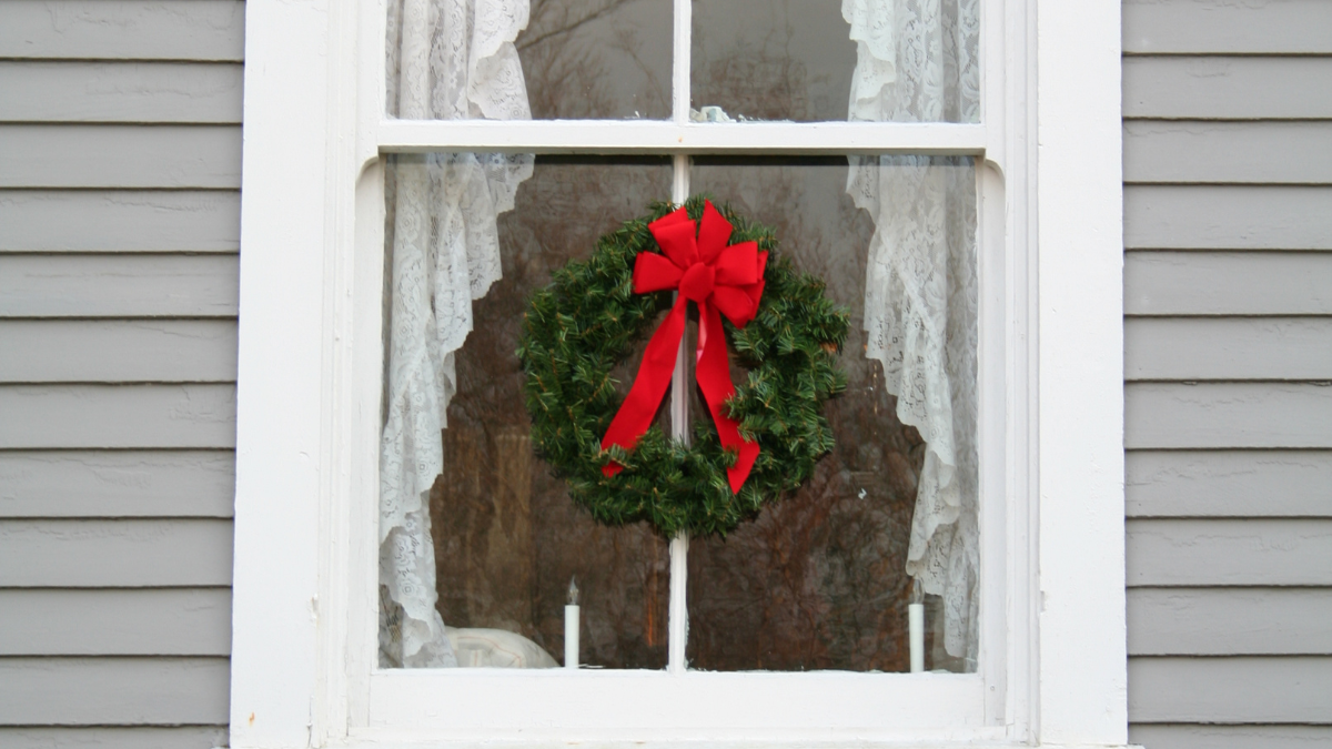A festive green Christmas wreath with a large red bow hangs in a white-framed window with lace