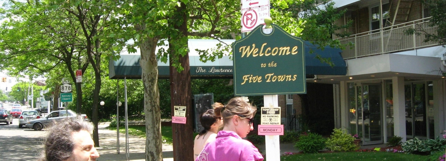 Green "Welcome to the Five Towns" sign on a tree-lined street with