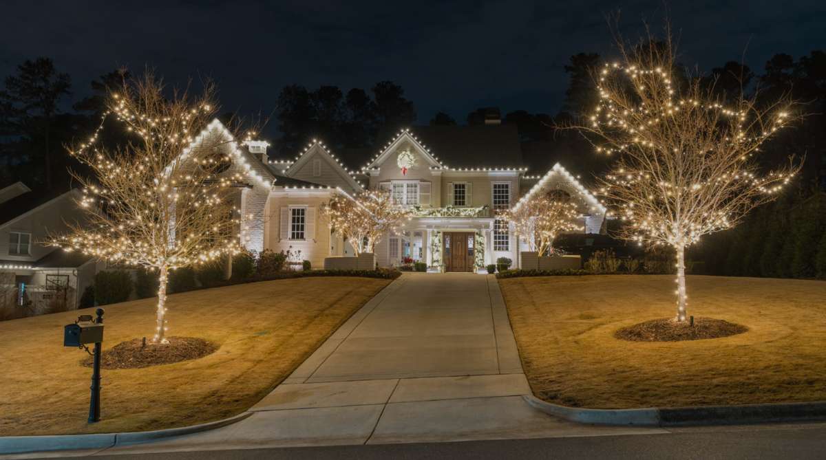 A grand house and bare trees are beautifully illuminated with warm white holiday lights at night.