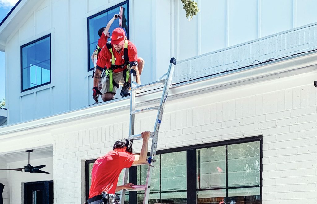 Two workers clean windows on a modern white house, one on the roof and another securing