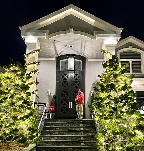 A man stands on the steps of a modern house decorated with festive Christmas lights at night