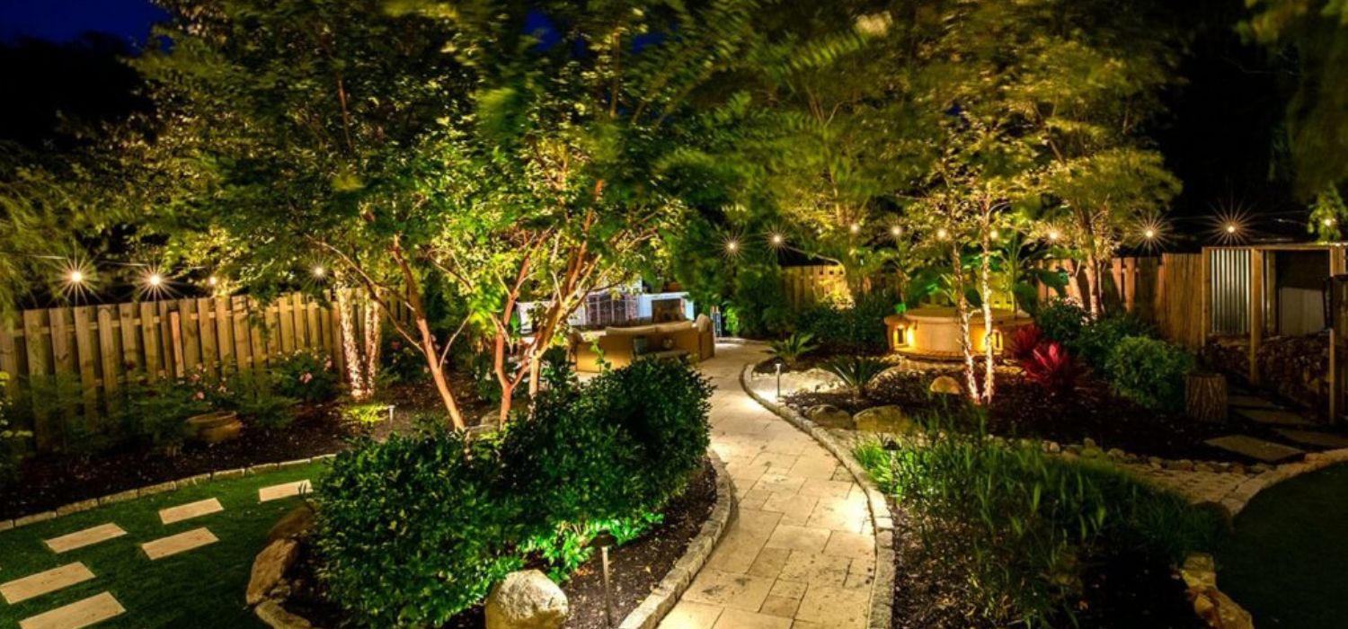 A beautifully illuminated backyard at night features a winding stone path, lush trees, outdoor seating