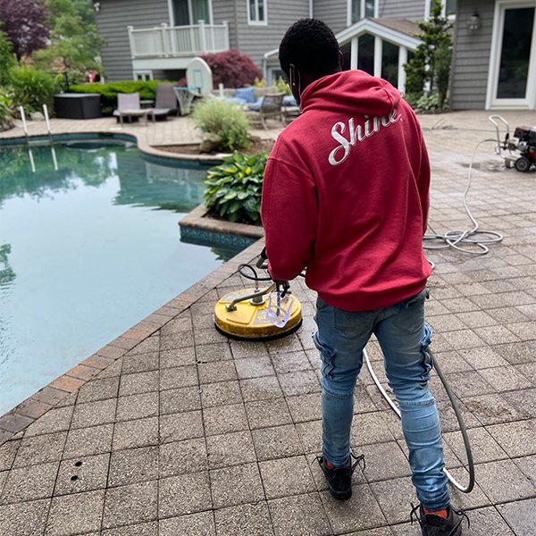 A person in a red 'Shine' hoodie power washes a paver patio next to