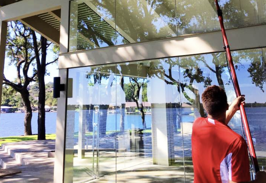 A person cleans large glass windows reflecting a scenic lake and lush trees. TITLE