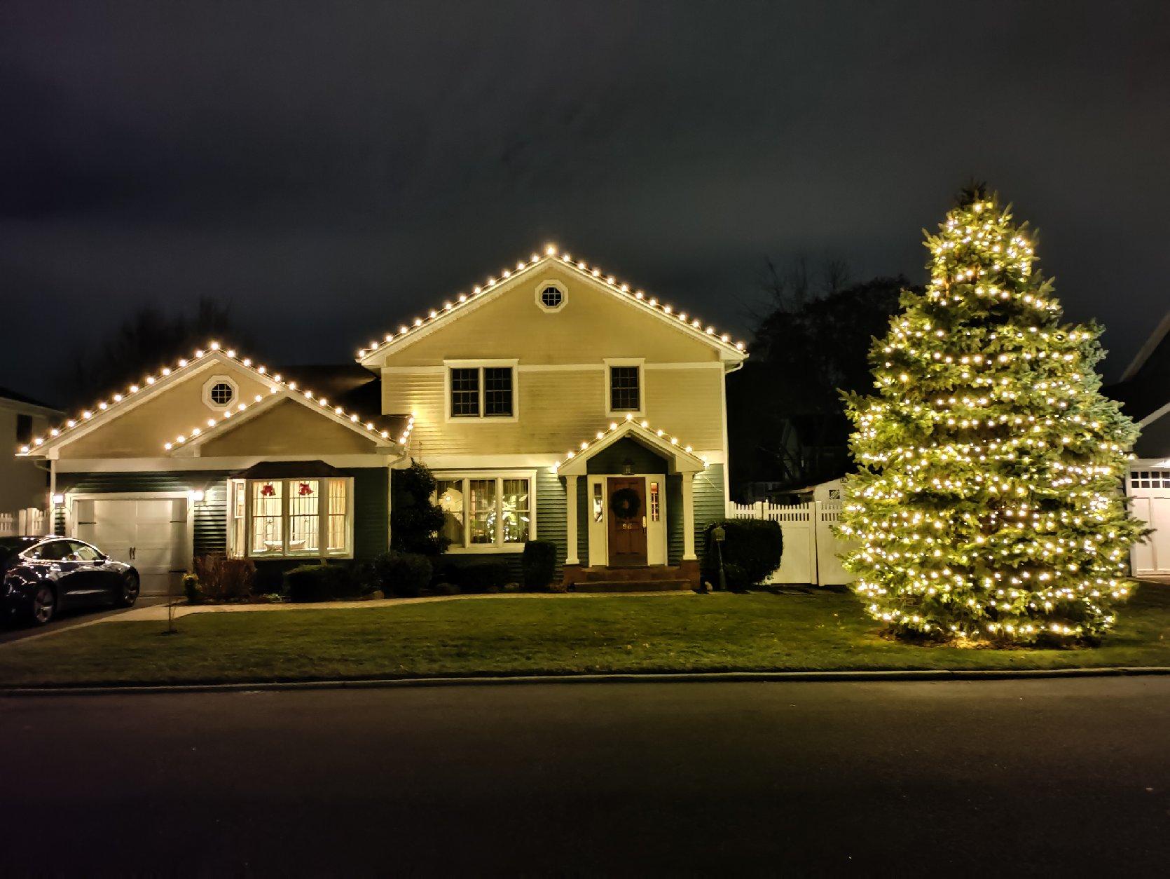 A house and large evergreen tree are brightly lit with warm white Christmas lights at night.