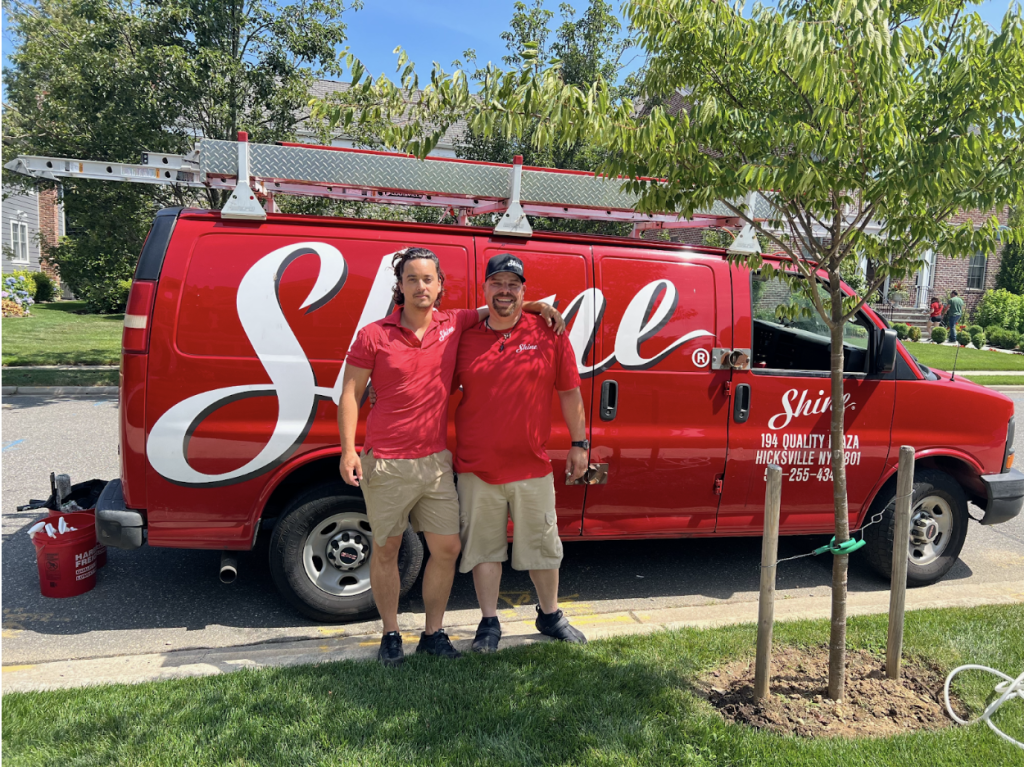 Two smiling men in red shirts stand proudly beside their red "Shine" service van with a