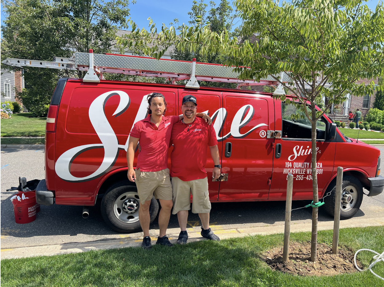 Two smiling men in red shirts stand proudly beside their red "Shine" service van with a
