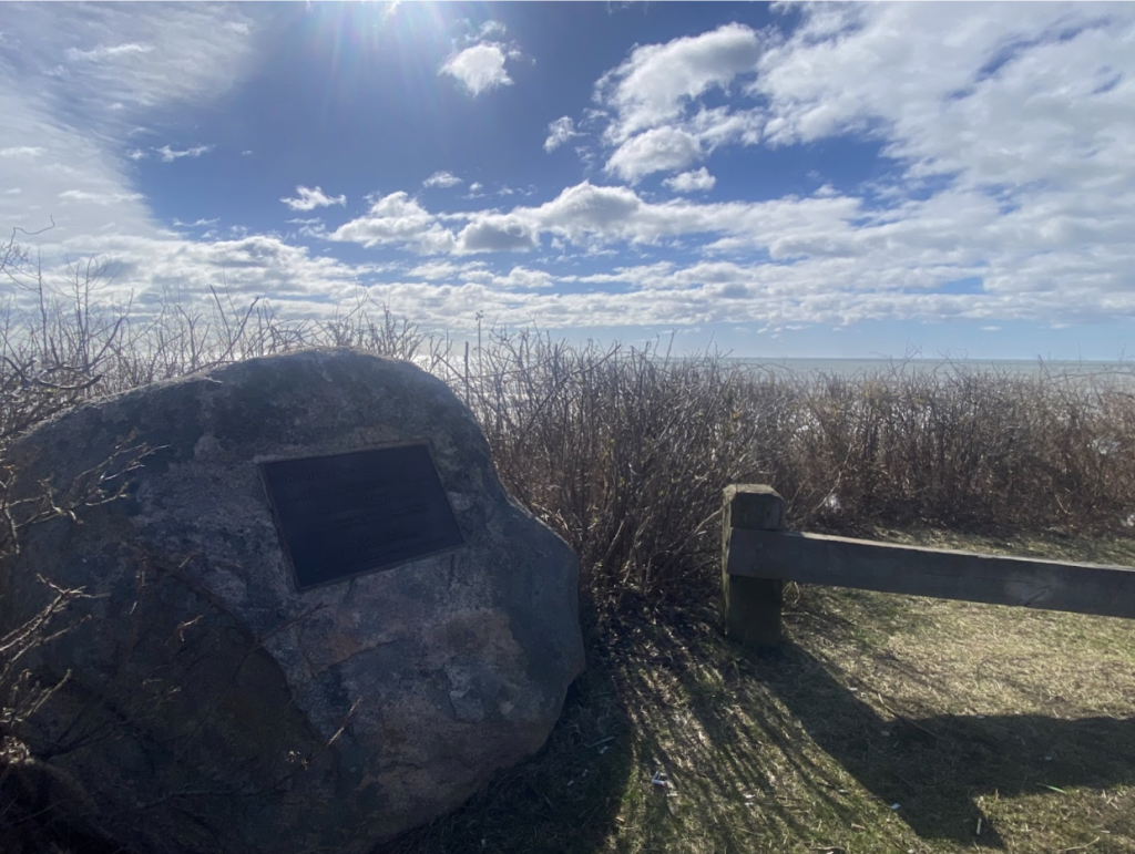 A large rock with a dark plaque sits among bare bushes overlooking calm water under a bright, cloudy