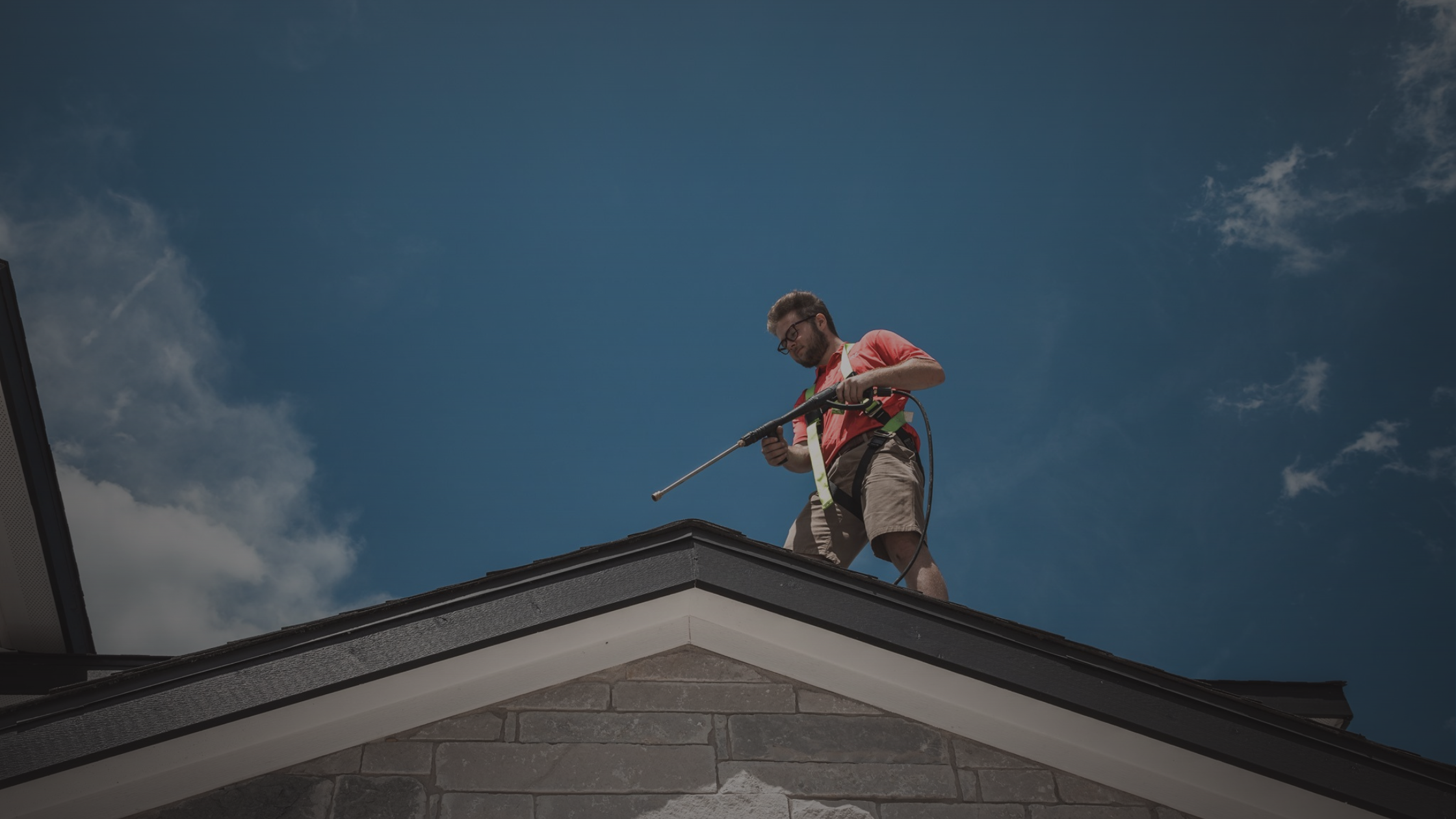 A man in a safety harness pressure washes a house roof under a blue sky.