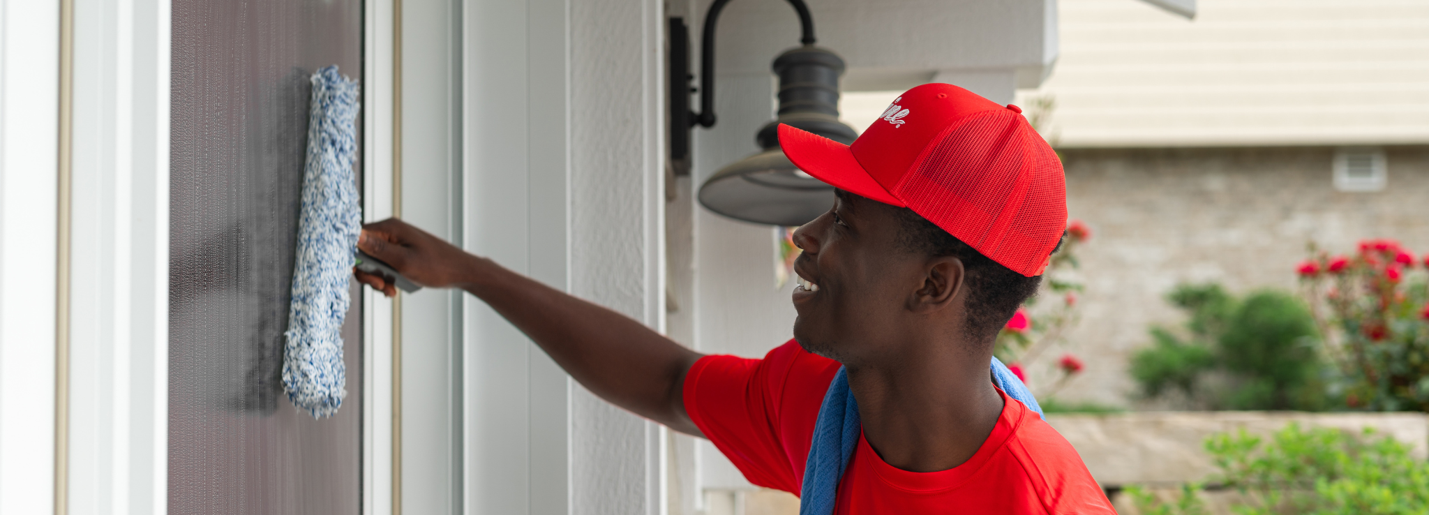 A smiling man in a red cap and shirt cleans a window with a blue scrubber. TITLE