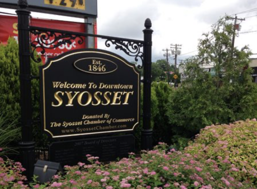 Ornate black and gold sign welcomes visitors to Downtown Syosset, surrounded by green bushes and