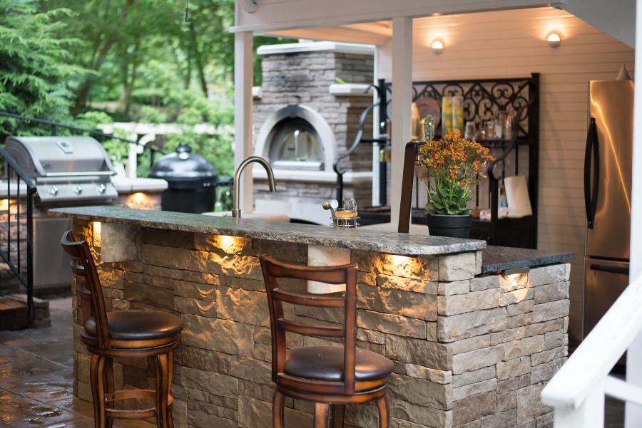 An inviting outdoor kitchen features a stone bar, grill, pizza oven, and stainless steel appliances.