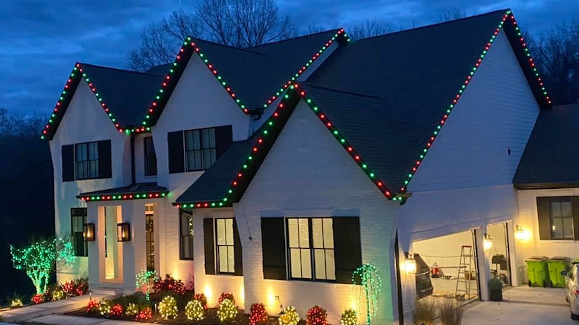 A modern white house is brightly lit with red and green Christmas lights along its roof and landscaping.