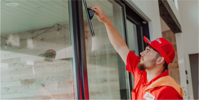 A bearded man in a red "Shine" uniform cleans a large residential window with