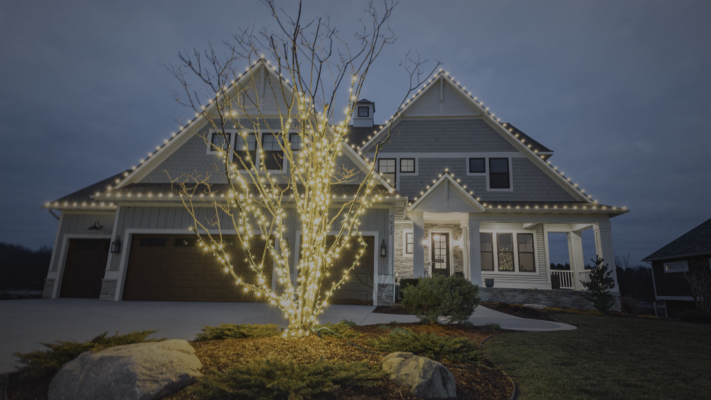 A modern house and its front yard tree are illuminated with warm white string lights at dusk