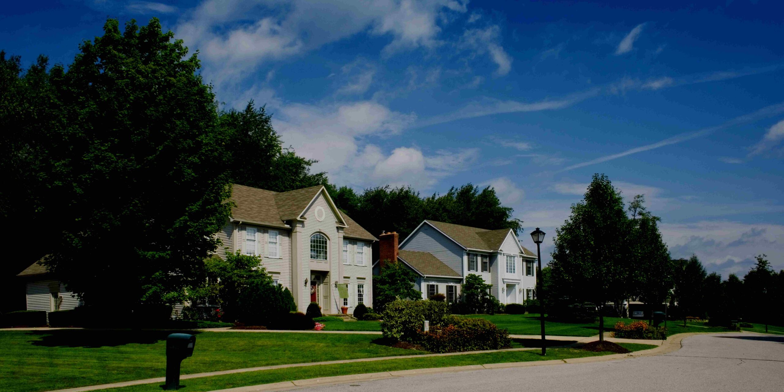 Suburban street with well-maintained houses, green lawns, and trees under a bright blue sky.