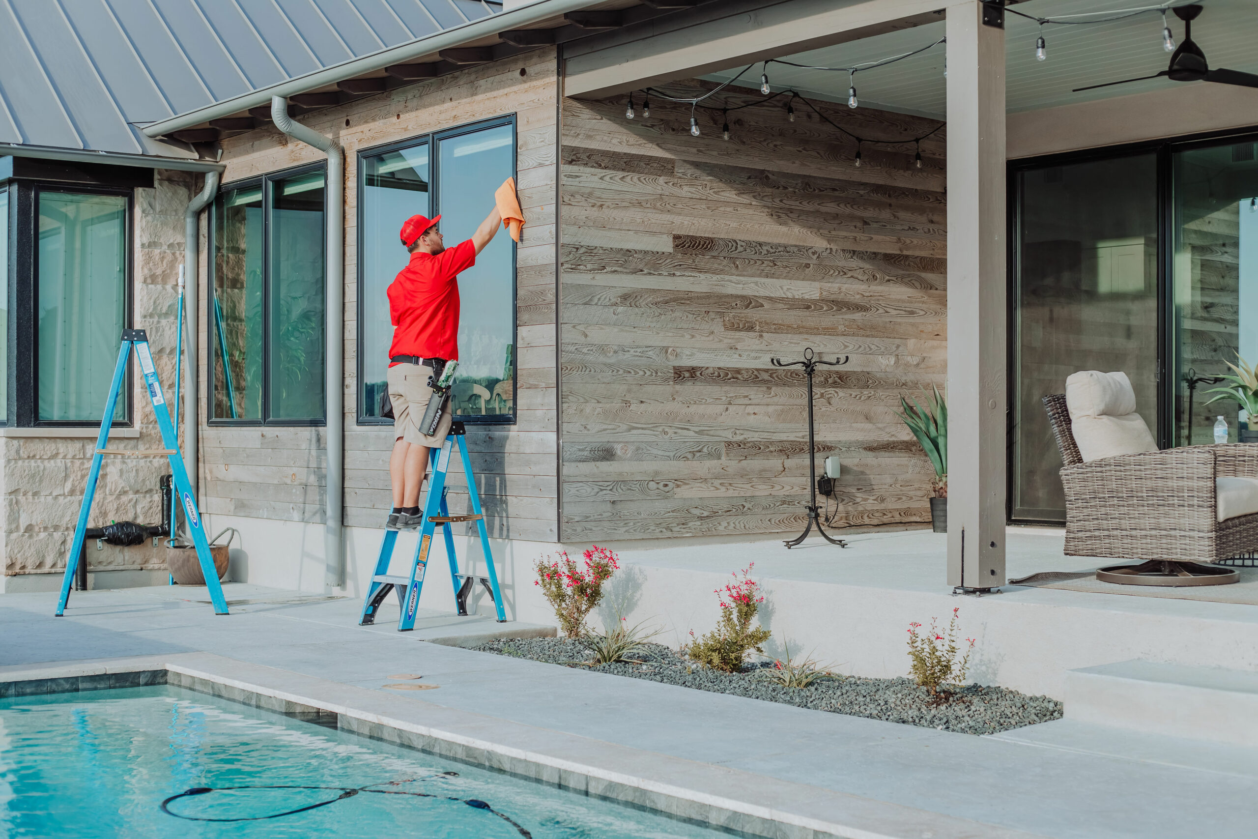 A man on a ladder cleans a large window of a modern house next to a swimming pool.