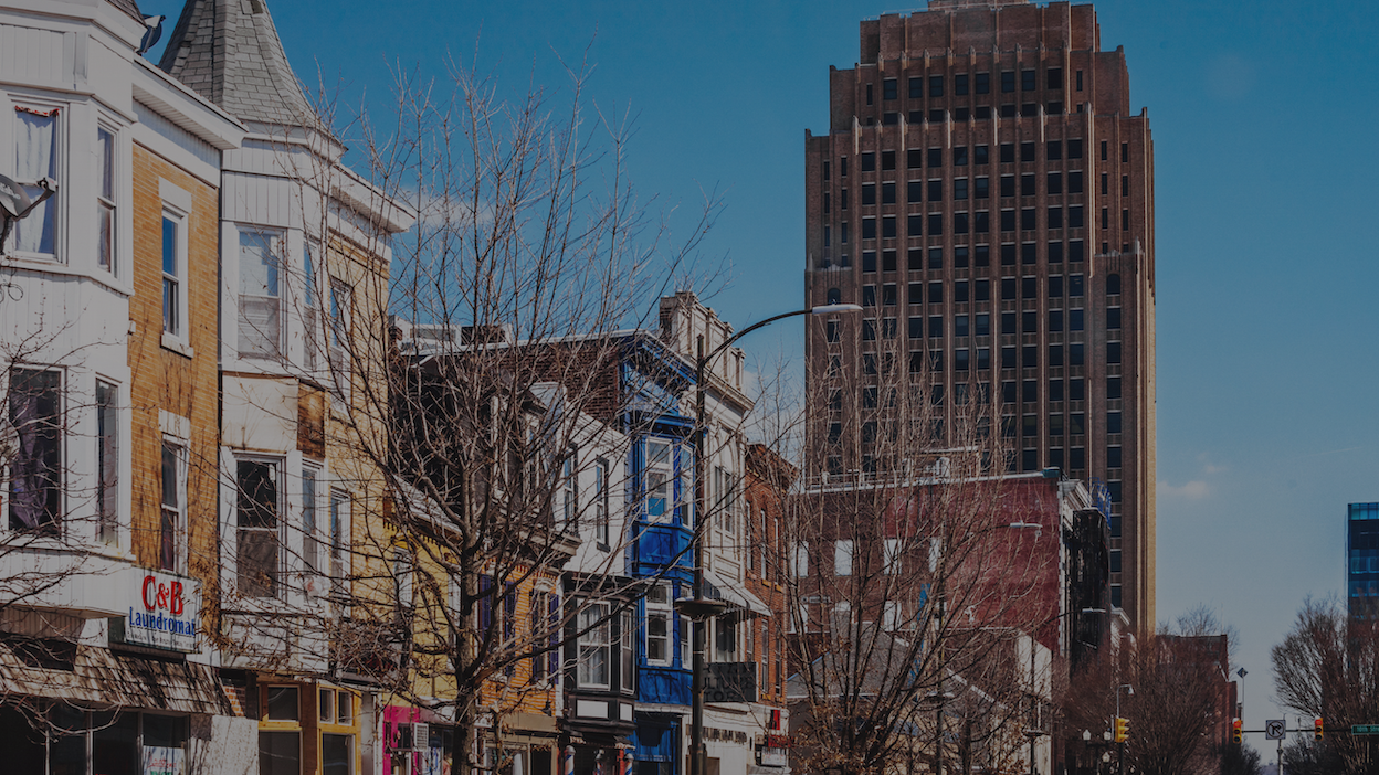 Colorful row houses line a street with bare trees, overlooked by a tall brown skyscraper
