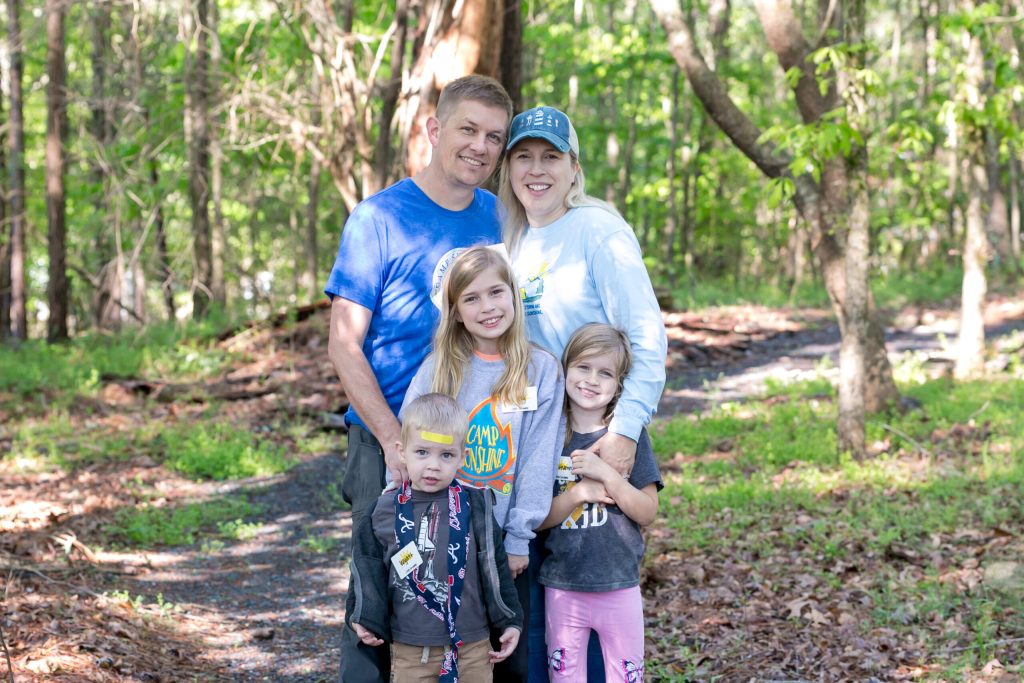A smiling family of four stands together in a sunny wooded area.