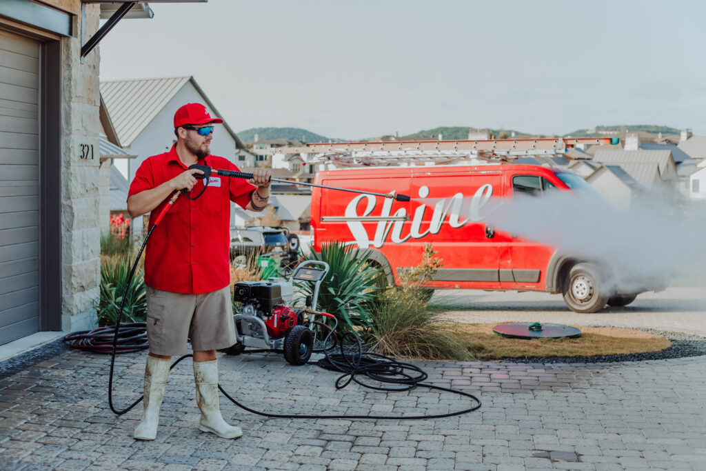 A professional in a red uniform pressure washes a driveway with a "Shine" service