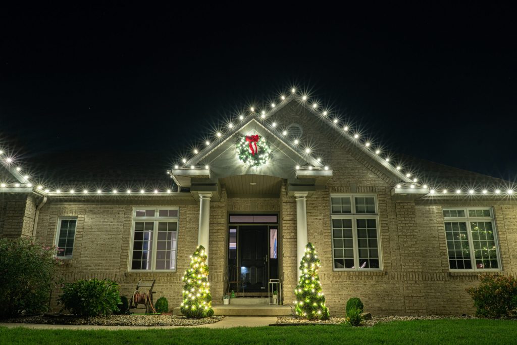 A brick house is beautifully decorated at night with white Christmas lights outlining the roof and a festive wreath