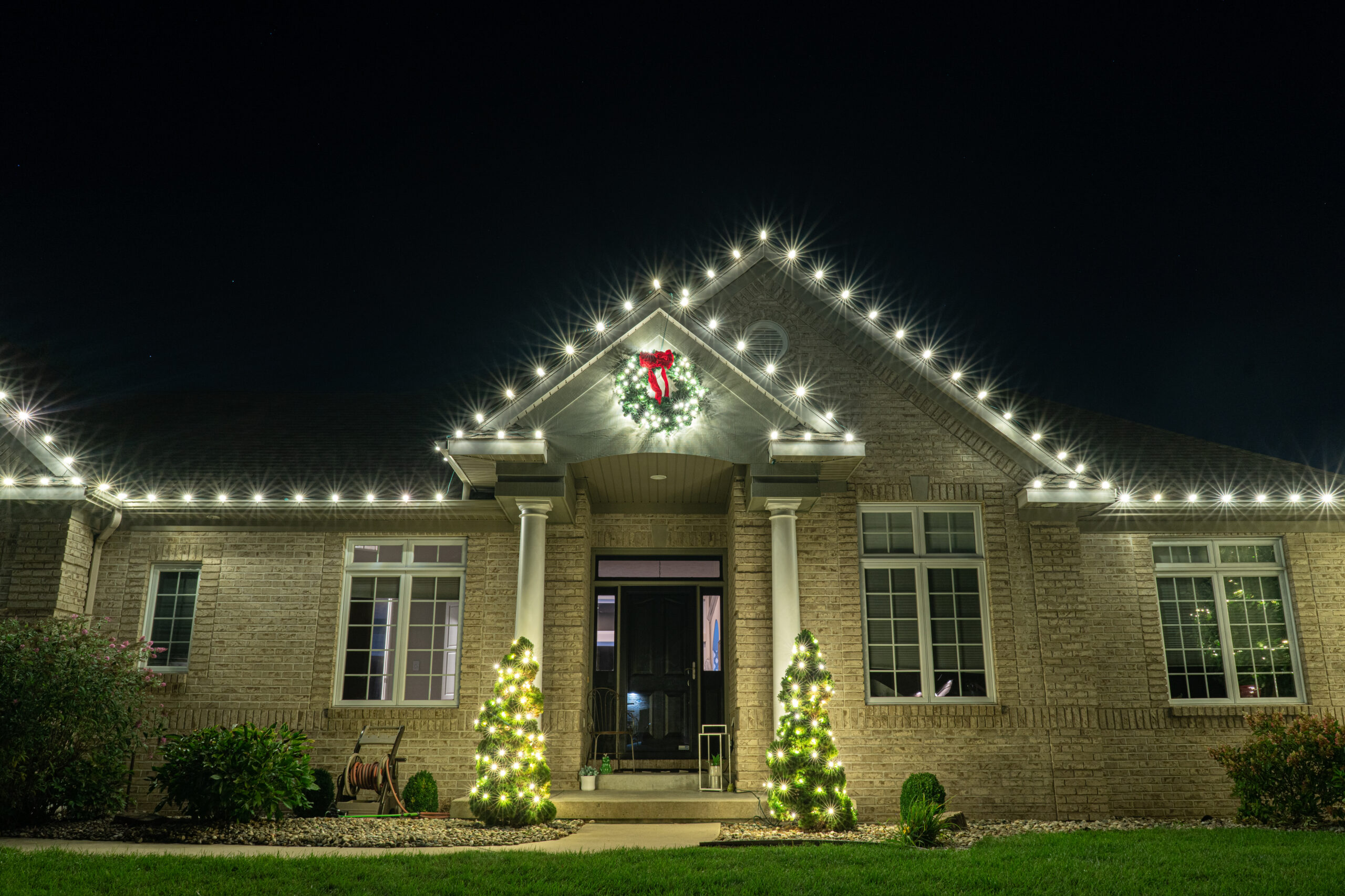 A brick house is beautifully decorated at night with white Christmas lights outlining the roof and a festive wreath