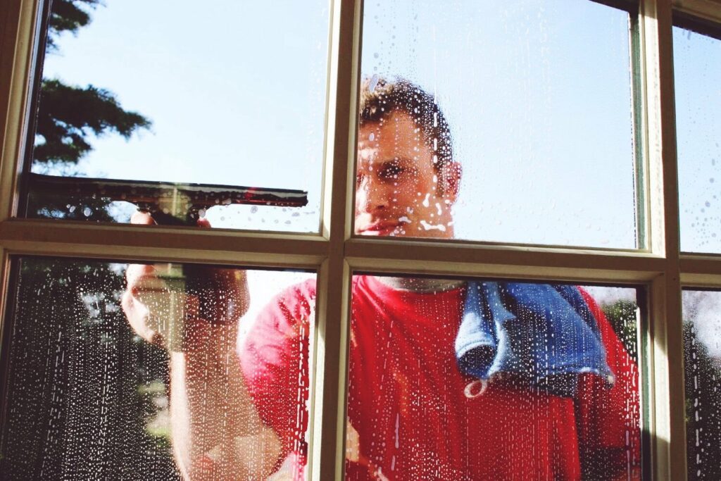 A man in a red shirt cleans a sudsy window with a squeegee.
