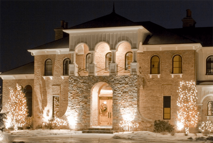 A grand brick house with a stone archway entrance is beautifully lit at night with festive lights. TITLE