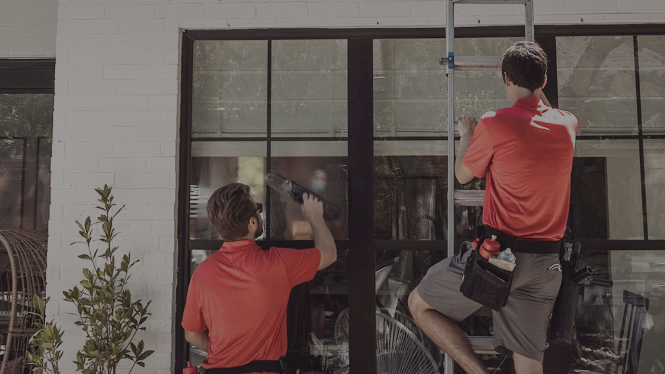 Two professional window cleaners in red shirts clean large windows on a white brick building.