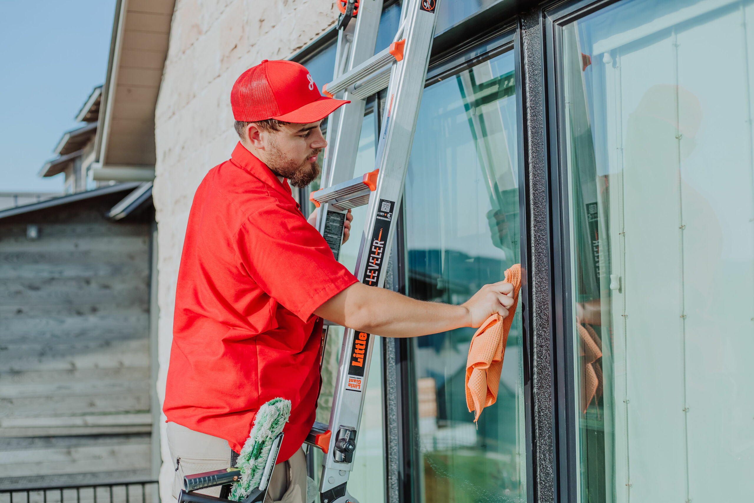 A man in a red shirt and cap cleans a large window from a ladder with an