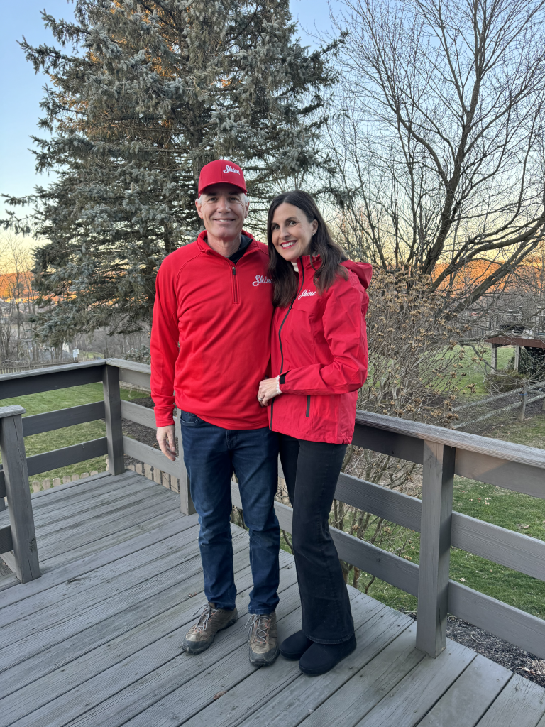 Happy couple wearing matching red "Shine" apparel poses on a wooden deck at sunset