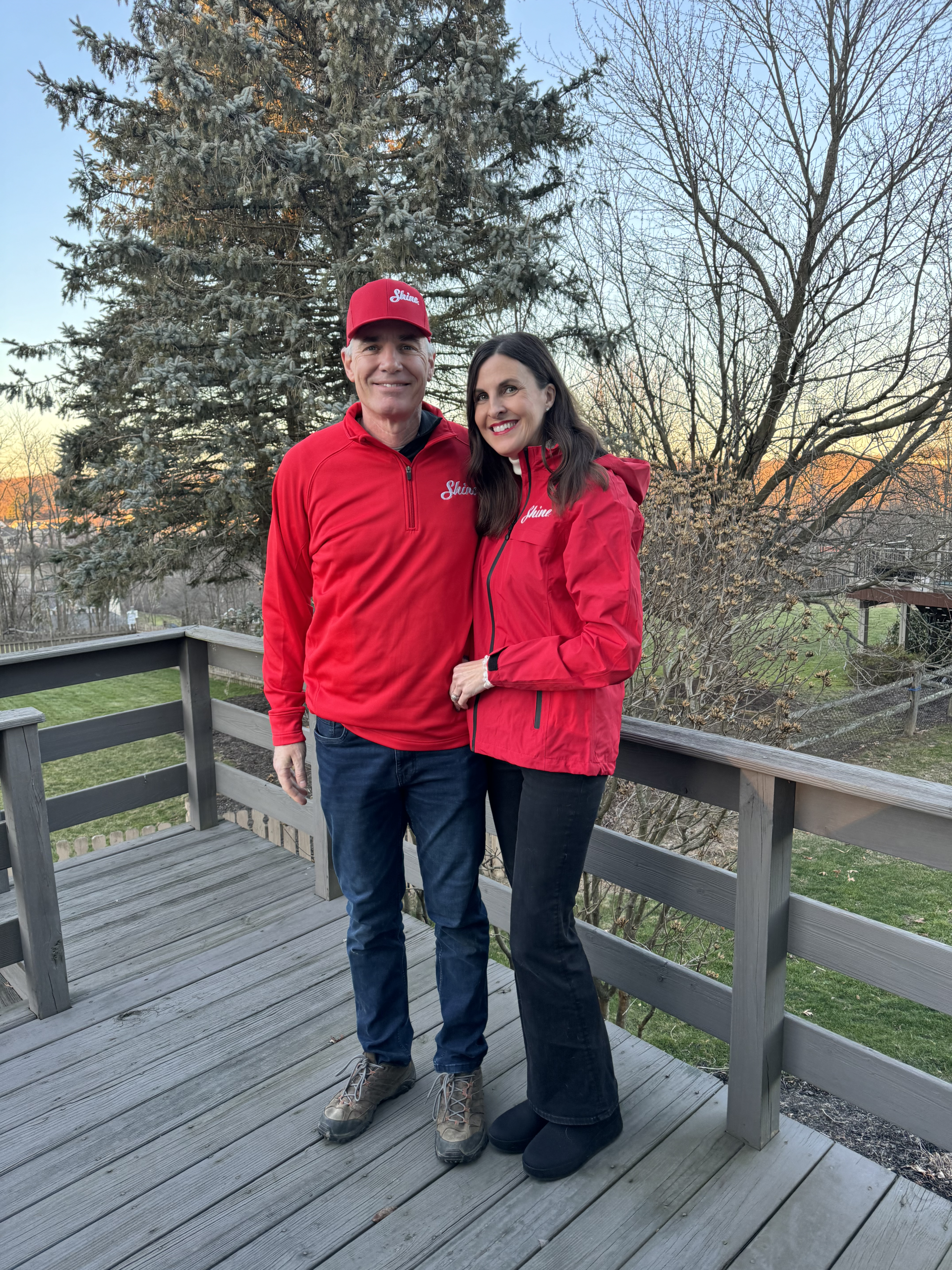 Happy couple wearing matching red "Shine" apparel poses on a wooden deck at sunset