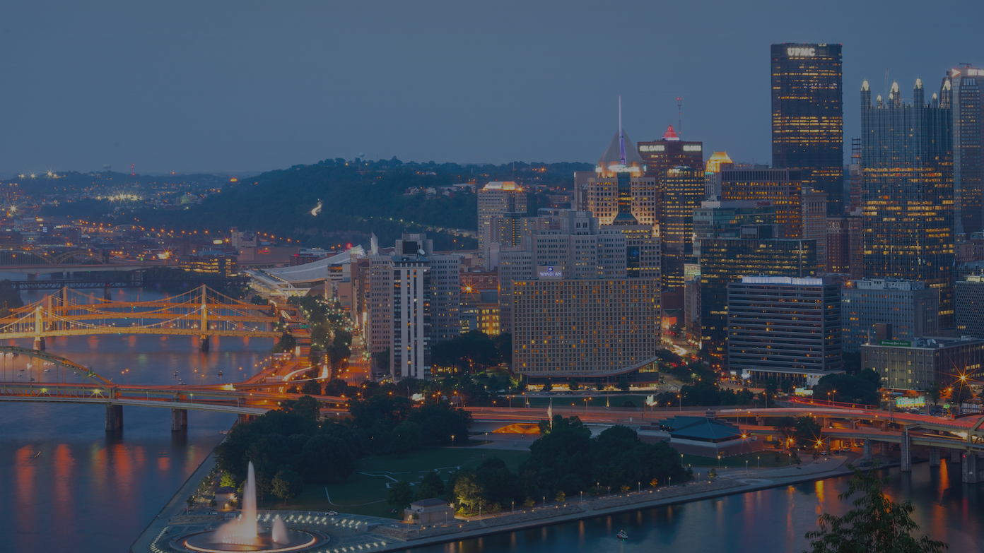 Twilight aerial view of Pittsburgh's illuminated city skyline, bridges, and river fountain.