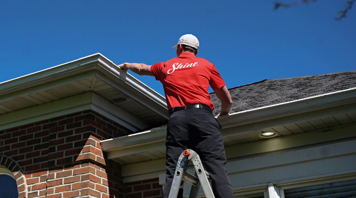 A service professional on a ladder cleans house gutters under a clear blue sky.