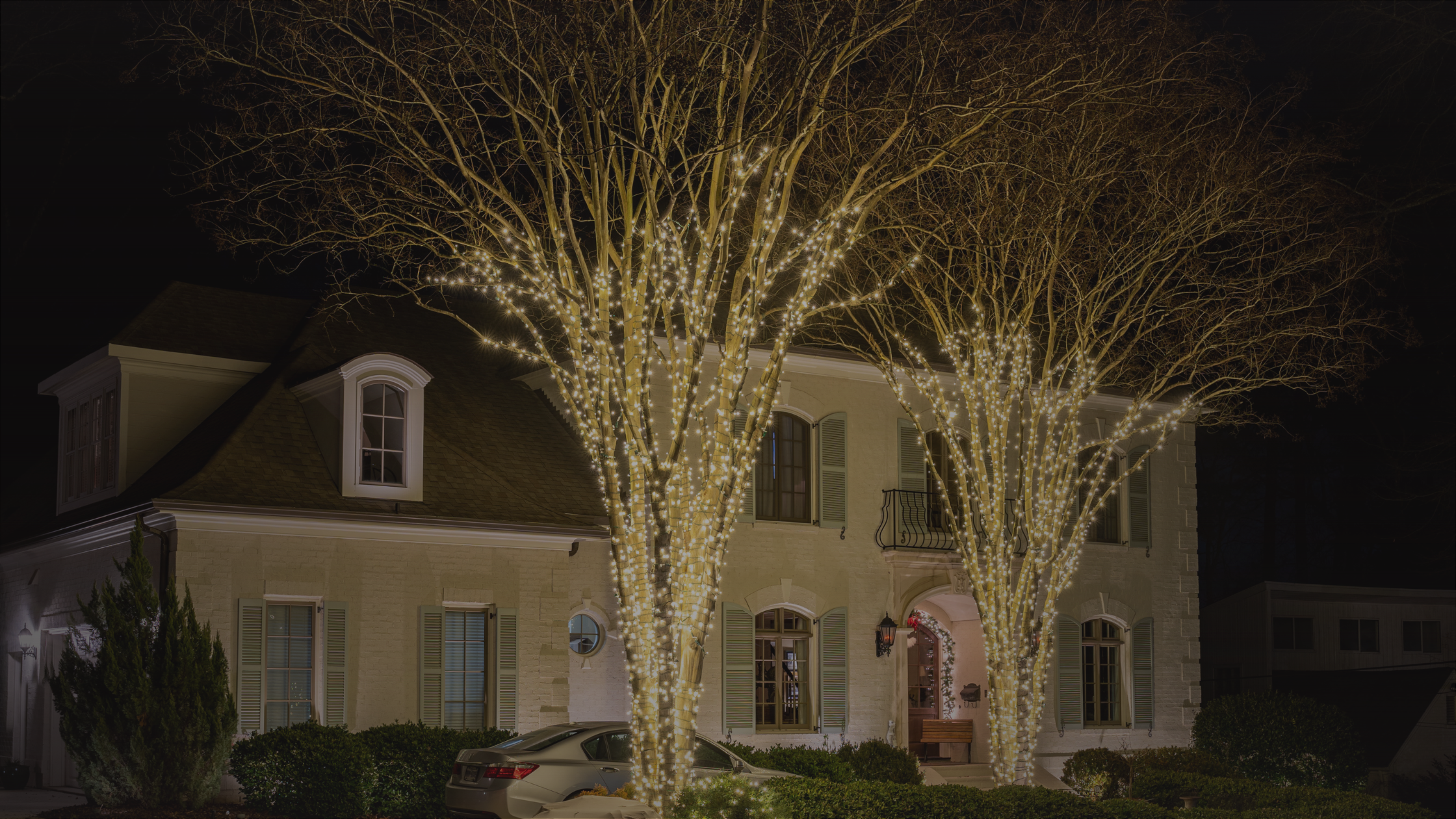 A stately home at night, framed by two bare trees intricately wrapped in glowing white lights