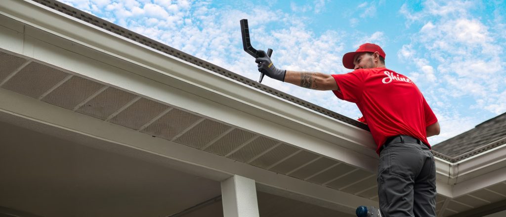 A worker in a red shirt and cap cleans a house gutter with a specialized tool.