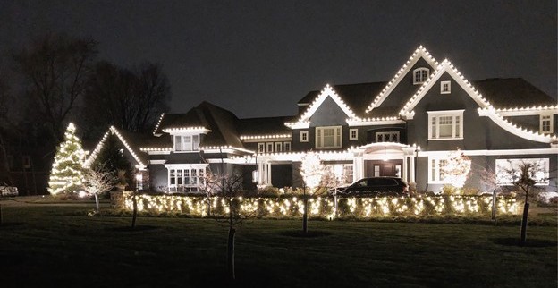 A large house and front yard are beautifully illuminated with warm white Christmas lights at night