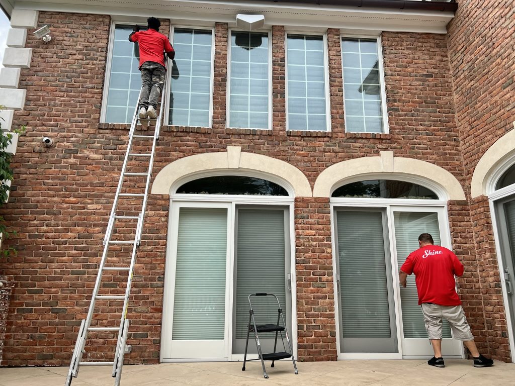 Professional window cleaners service a multi-story brick home, one on a ladder and one on the ground.