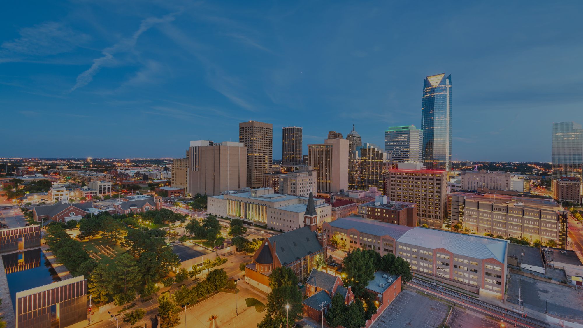 A city skyline at twilight features a tall skyscraper, illuminated buildings, and a reflective park.