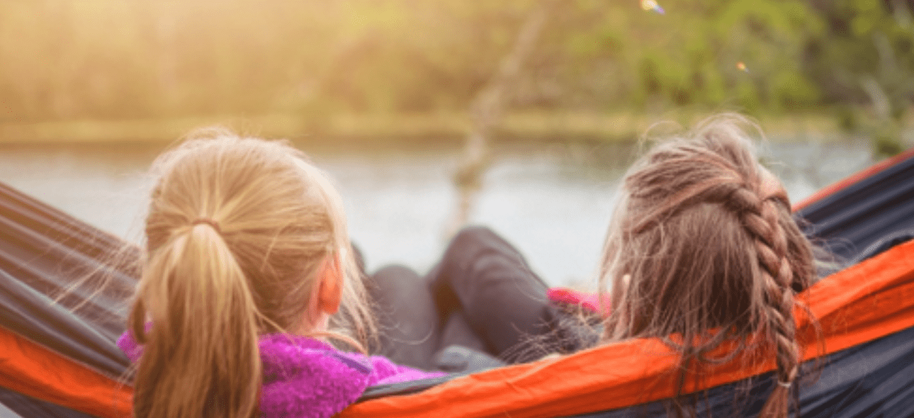 Two girls relax in a hammock, looking out at a sunny lake and trees. TITLE: