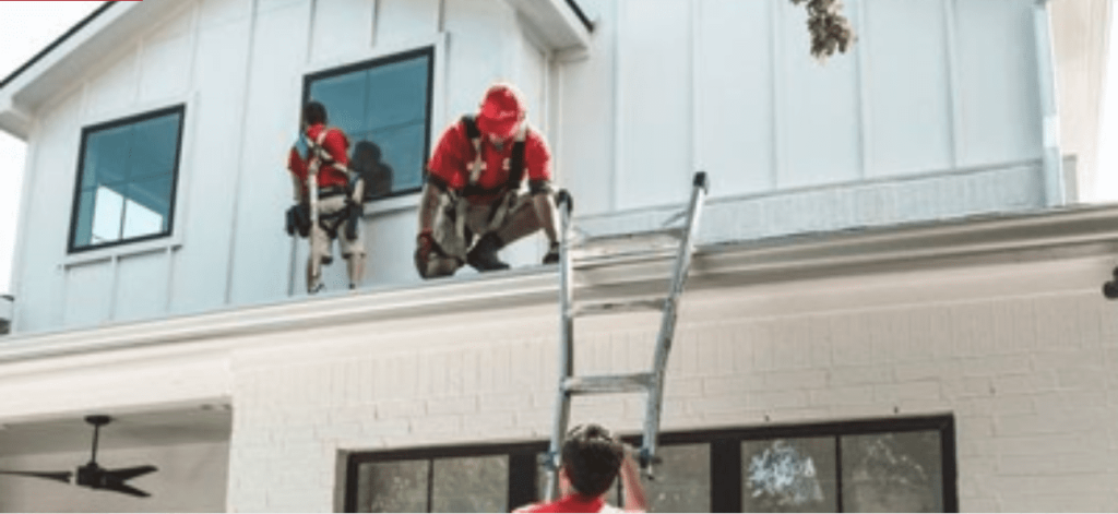 Roofers in red safety gear work on a white house roof with a ladder.