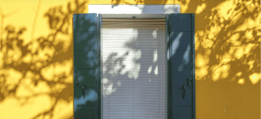 A window with green shutters and blinds is bathed in sunlight, casting tree shadows on a yellow wall.