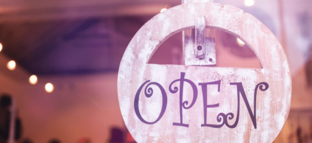 A rustic wooden "OPEN" sign hangs in a warm, inviting setting with blurred string lights.