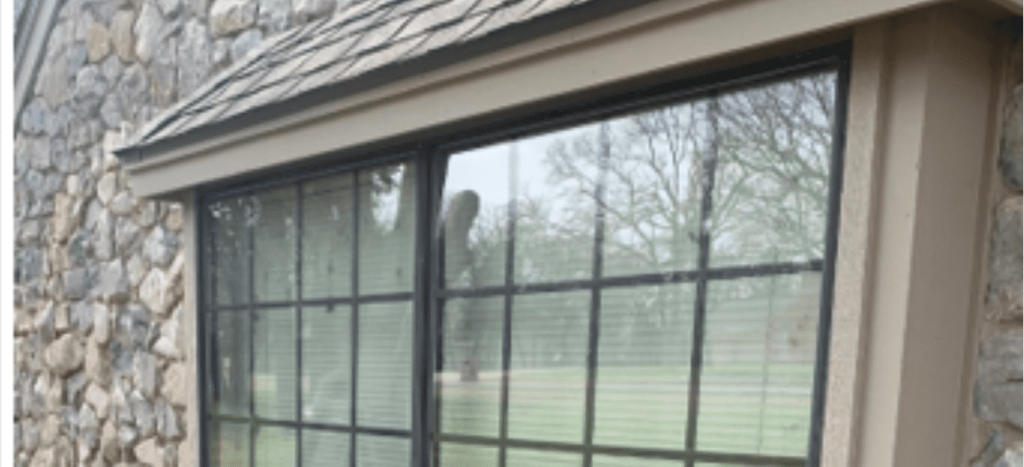 A stone house features a large window with dark frames, reflecting bare trees and a grey